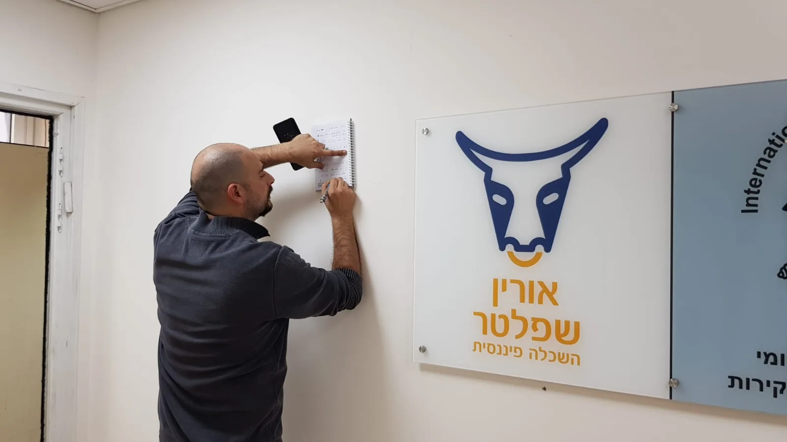 סטודנט תולה מחברת על קיר בכיתה במכללת אורין שפלטר, ליד שלט עם לוגו המכללה